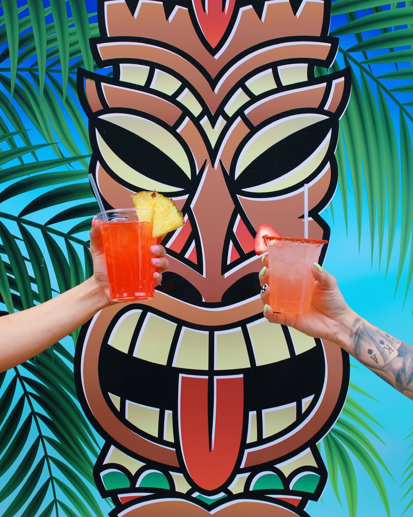 Two frozen tiki drinks raised in front of the Tipsy Tiki mural